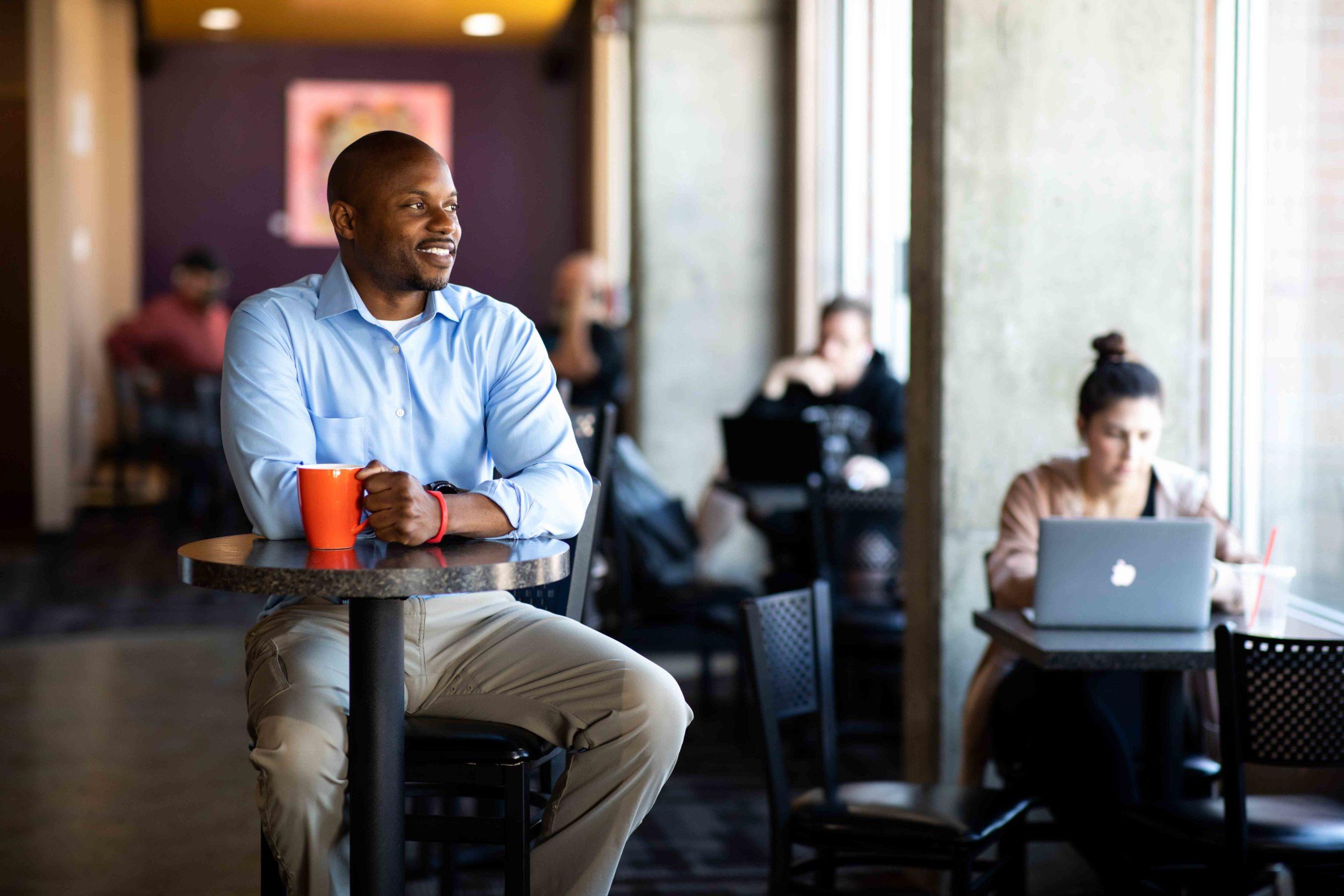 密歇根州立大学丹佛 Grad Ryan Cobbins sitting in a coffee shop with a mug in his hand, looking out the window.