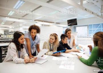 老师带着一群大学生，在实验室的教室里. 老师正在考虑一个学生的工作，心情轻松而积极. 其他同学正在互相讨论事情. 这是一个真实的教学场景，坦率的表达. 这是一个多民族的妇女群体. 背景中有一块写着数学公式的白板. 所有的女士都戴着身份牌.