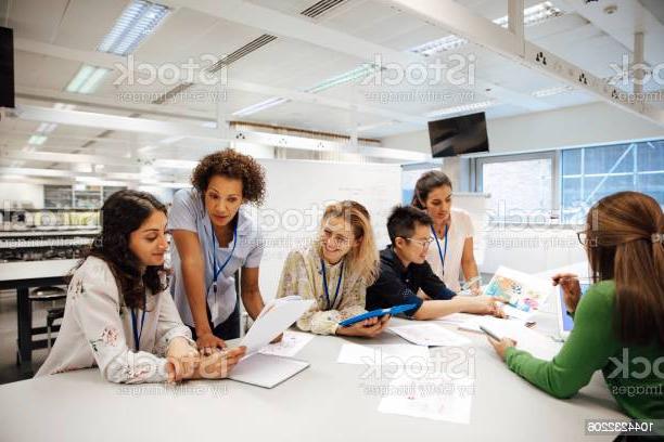 老师带着一群大学生，在实验室的教室里. 老师正在考虑一个学生的工作，心情轻松而积极. 其他同学正在互相讨论事情. 这是一个真实的教学场景，坦率的表达. 这是一个多民族的妇女群体. 背景中有一块写着数学公式的白板. 所有的女士都戴着身份牌.