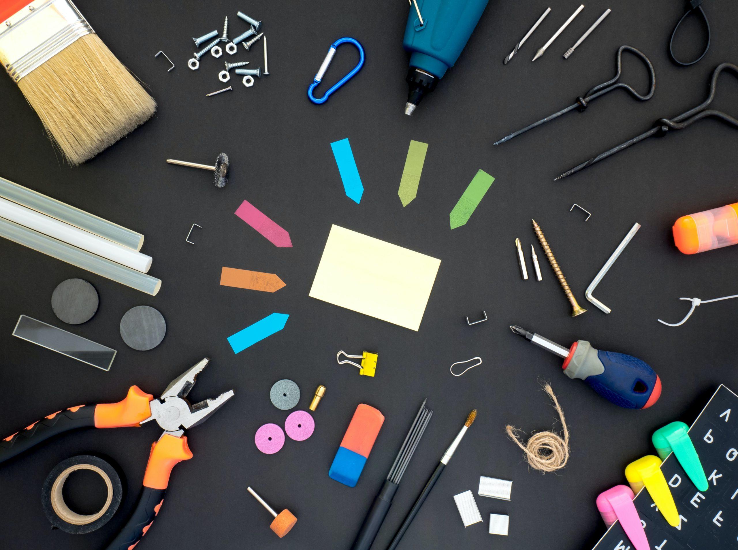 各种各样的工具放在黑色的表面上，包括便利贴, 螺丝刀, 扳手, 磁带, 画笔, 笔用, 胶棒, 斯台普斯, 磁铁, 和回形针.
