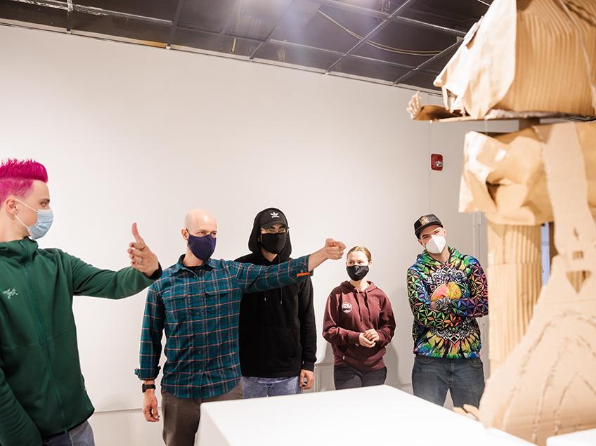 迈克尔·伯恩哈特(Michael Bernhardt)教授在199号画廊与他的学生一起进行评论. 通过他们的手势, 他们正在讨论前景中基座上的纸板雕塑的某些角度. 一个学生有一头电粉色的头发.