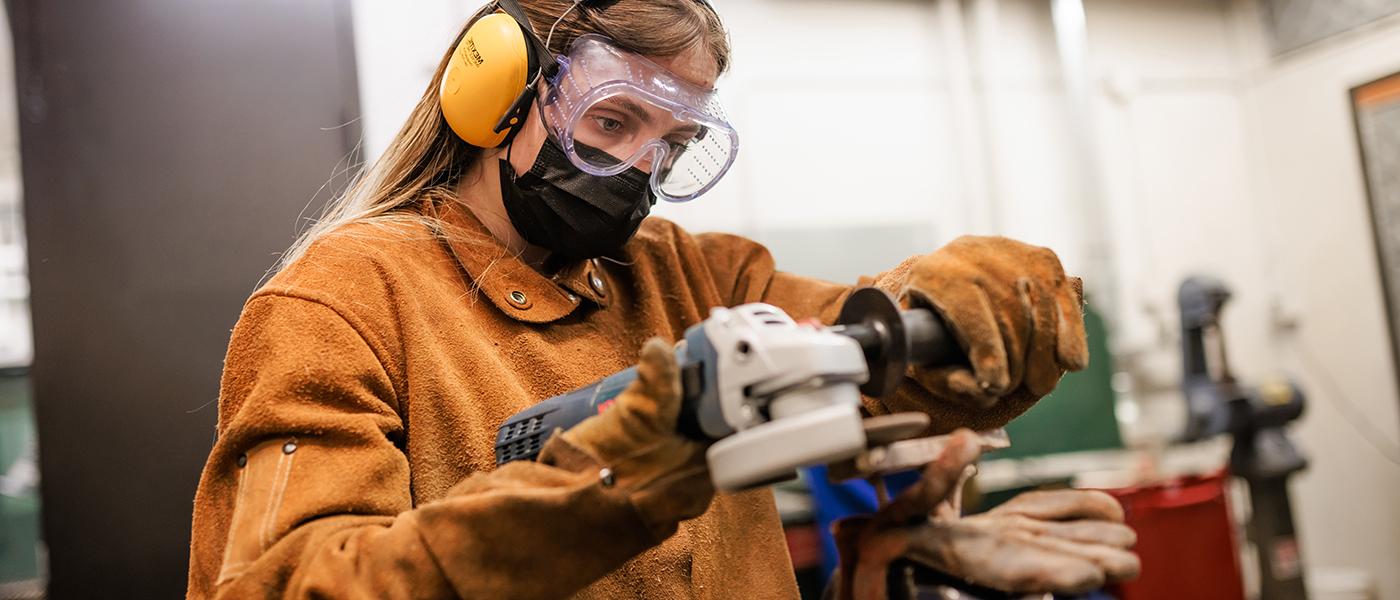 学生穿戴防护装备，包括护目镜, 耳机, 面具, 手套在雕塑工作室工作.