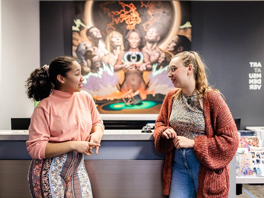 两个学生站在艺术系的前台聊天. 在他们身后是一幅画，画中有代表地球上许多民族的不同群体的人. 他们拿着一个中央浮动的球体和立方体，摆出沉思和沉思的姿势.