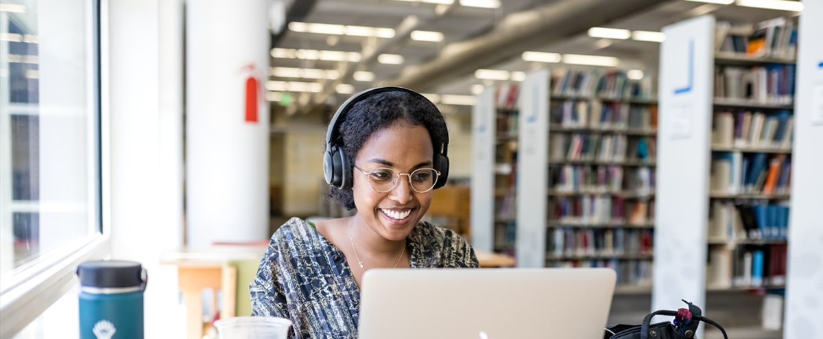 一个学生戴着耳机坐在图书馆的电脑前，面带微笑.