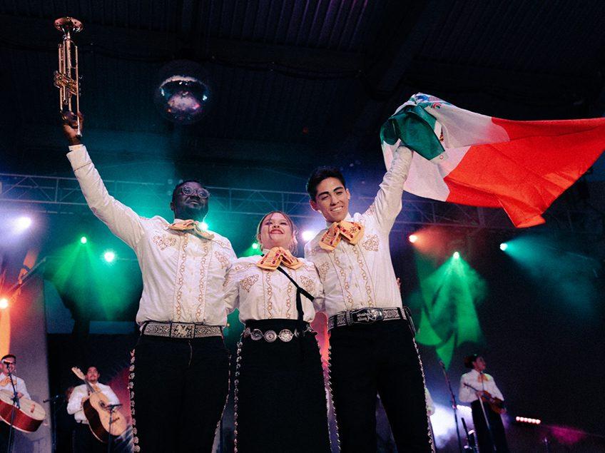 三名墨西哥流浪乐队表演者手持小号和墨西哥国旗向观众挥手致意